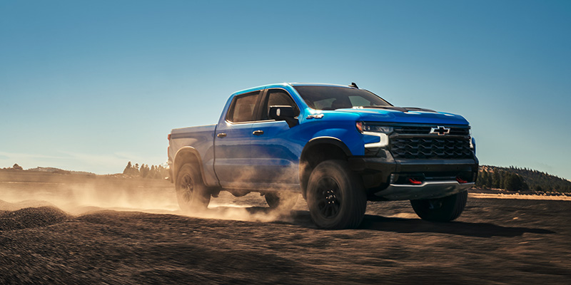 A blue 2025 Chevy Silverado 1500 driving through rocky terrain on a sunny day with blue skies overhead
