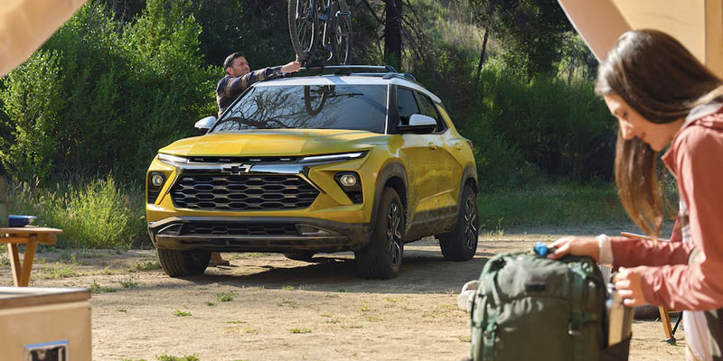 A yellow Chevy Trailblazer parked at a campsite on a sunny day with a man securing a bike to the roof rack of the vehicle