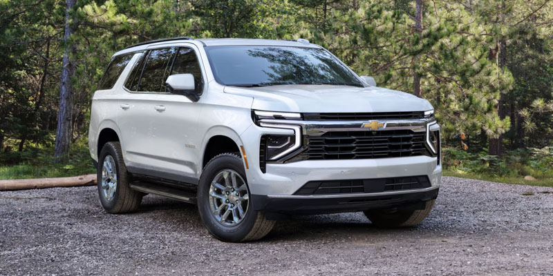 Front passenger view of a white 2025 Chevy Tahoe parked on gravel on a sunny day with trees in the background